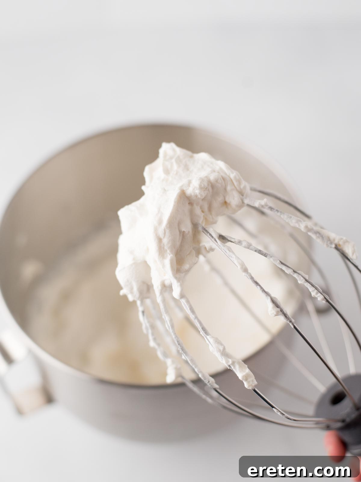 Easy Homemade Smores Ice Cream 8 A close-up of a metal whisk covered in freshly whipped cream, held above a mixing bowl filled with more whipped cream. The background is softly blurred.