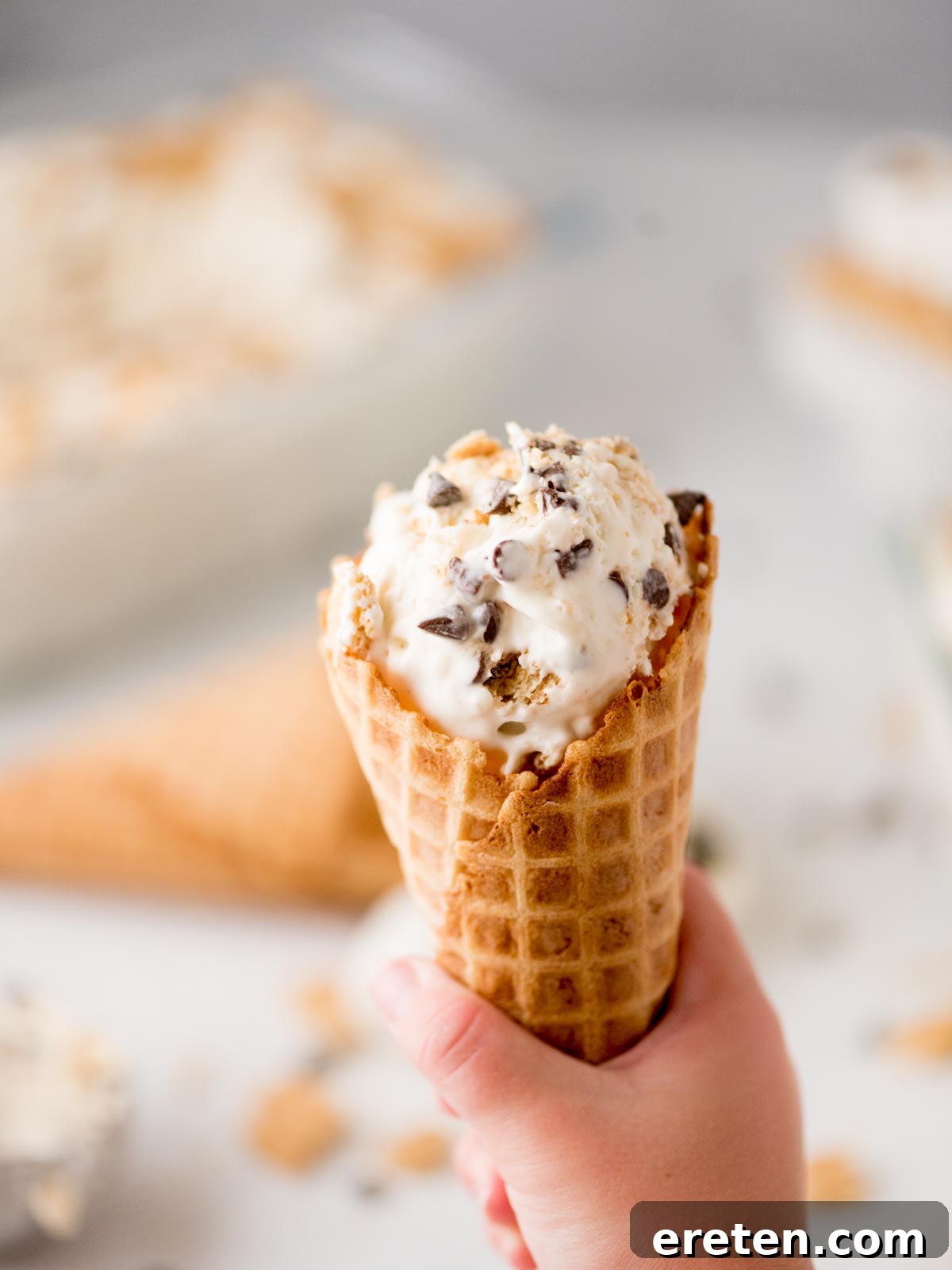 Easy Homemade Smores Ice Cream 9 A hand holding a waffle cone filled with ice cream containing chocolate chunks and cookie pieces, with another empty cone and blurred ice cream in the background.