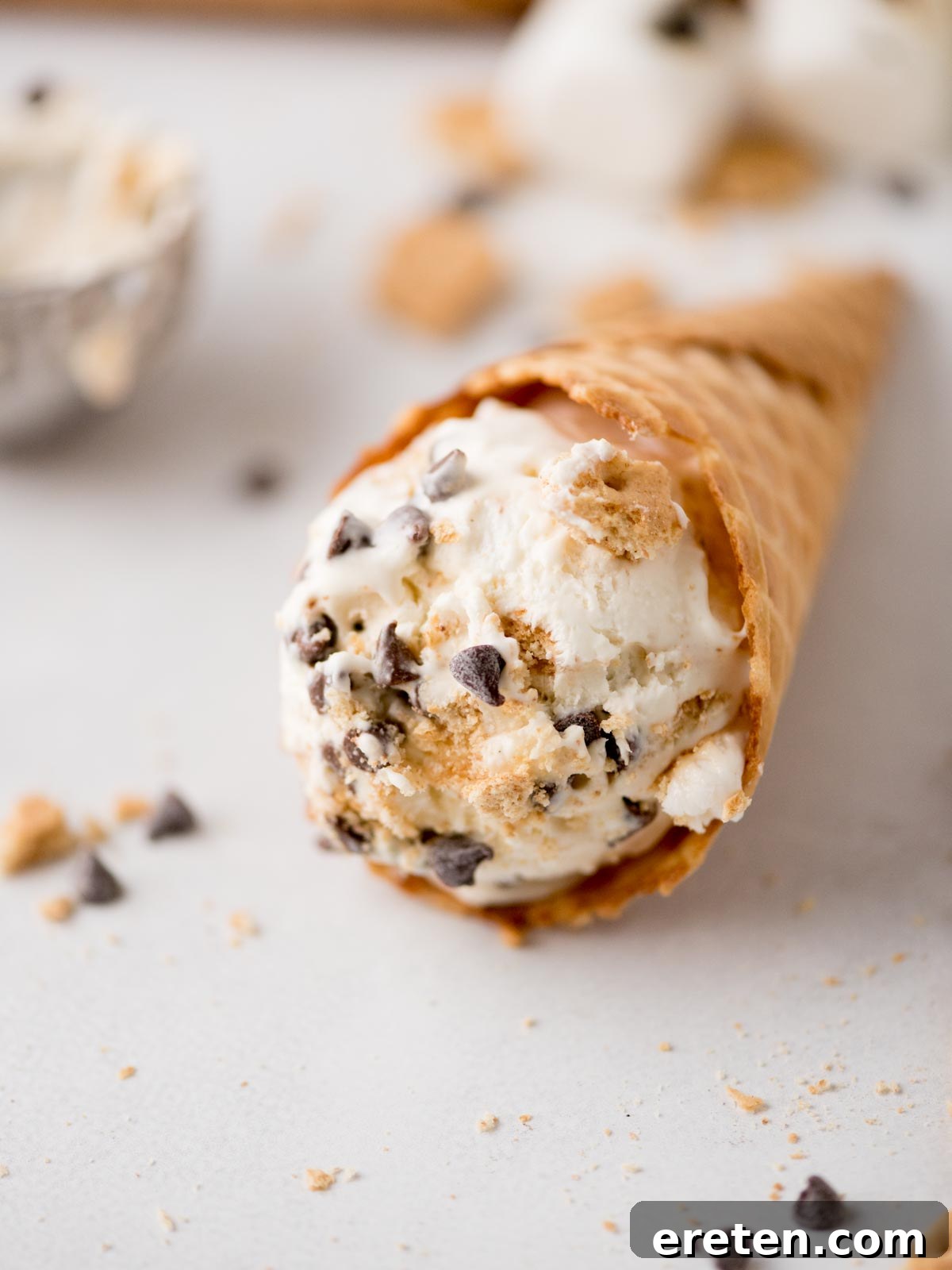 Easy Homemade Smores Ice Cream 10 A waffle cone filled with a scoop of ice cream containing chocolate chips and cookie crumbs, lying on its side with some chocolate chips and crumbs scattered nearby on a light surface.