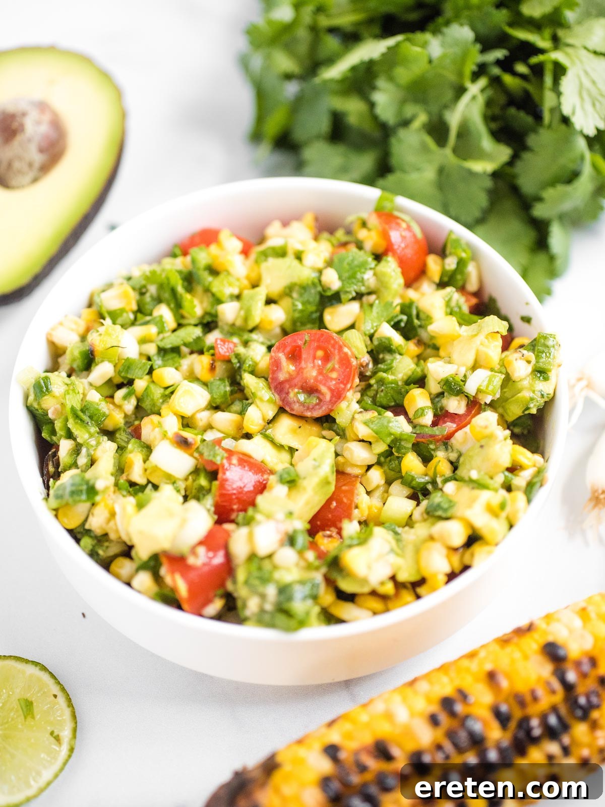 Vibrant summer corn salad in a white bowl, bursting with fresh corn, tomatoes, and avocado, garnished with fresh cilantro.