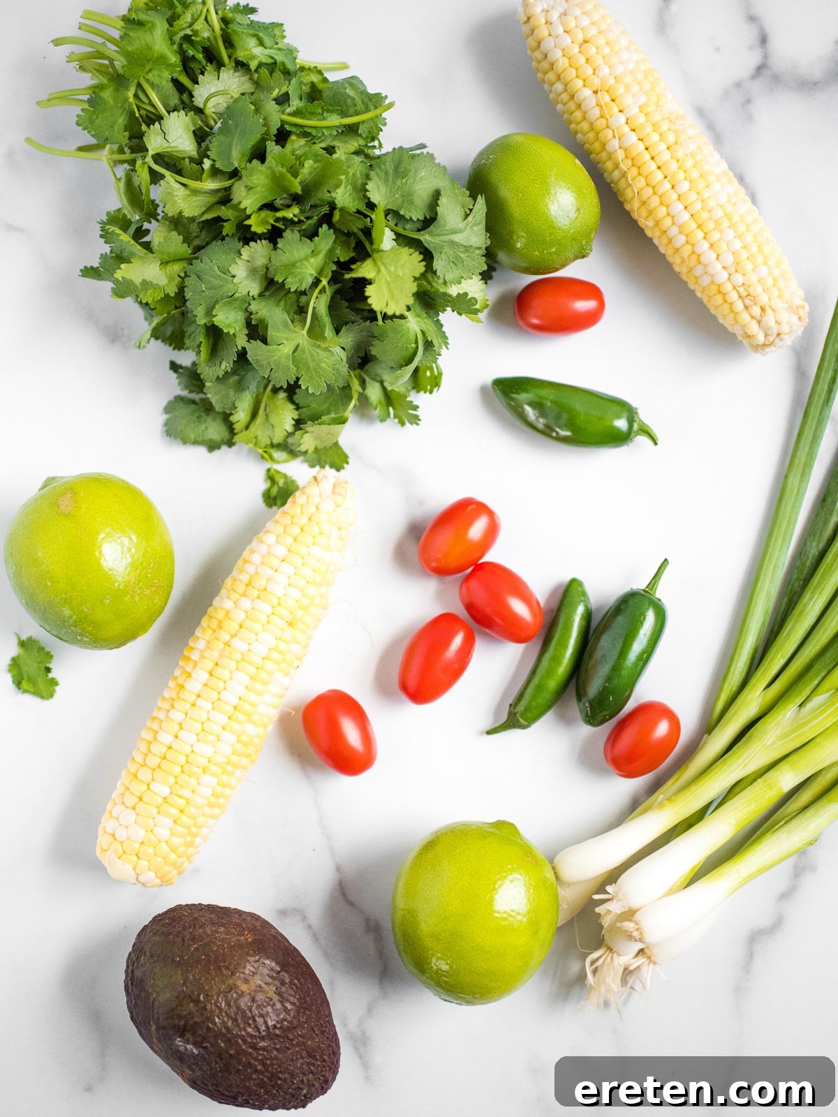 Fresh ingredients laid out on a wooden board for summer corn salad, including vibrant grape tomatoes, creamy avocado, grilled corn, fresh cilantro, sliced green onion, garlic cloves, and a zesty jalapeno.
