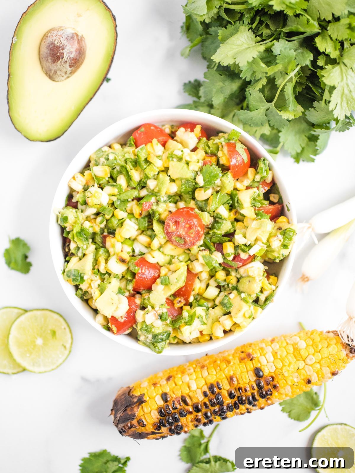 A bowl of vibrant summer corn salad, garnished with fresh cilantro, sits next to additional fresh ingredients like lime and a cutting board with more tomatoes.