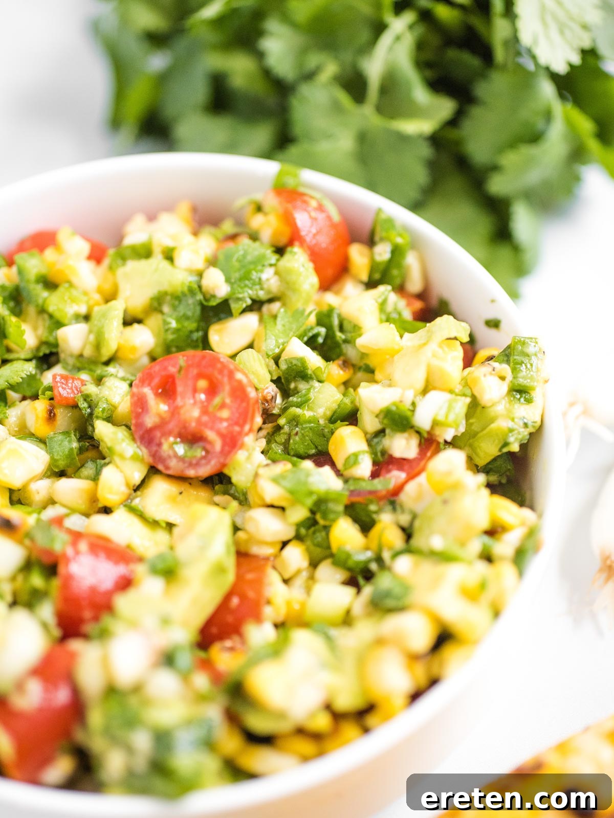 A close-up of a refreshing summer corn salad in a white bowl, highlighting the vibrant colors and textures of perfectly grilled corn, ripe tomatoes, and creamy avocado, ready to be enjoyed.