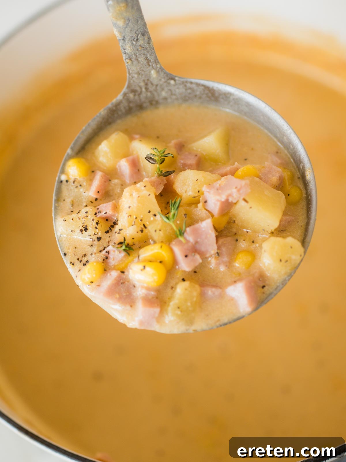 A ladle serving a generous portion of creamy ham and potato corn chowder from a large pot.