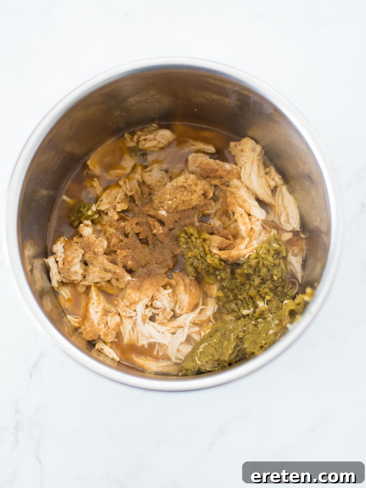A mix of aromatic spices and chopped green chiles being stirred into the shredded chicken and broth in the Instant Pot.