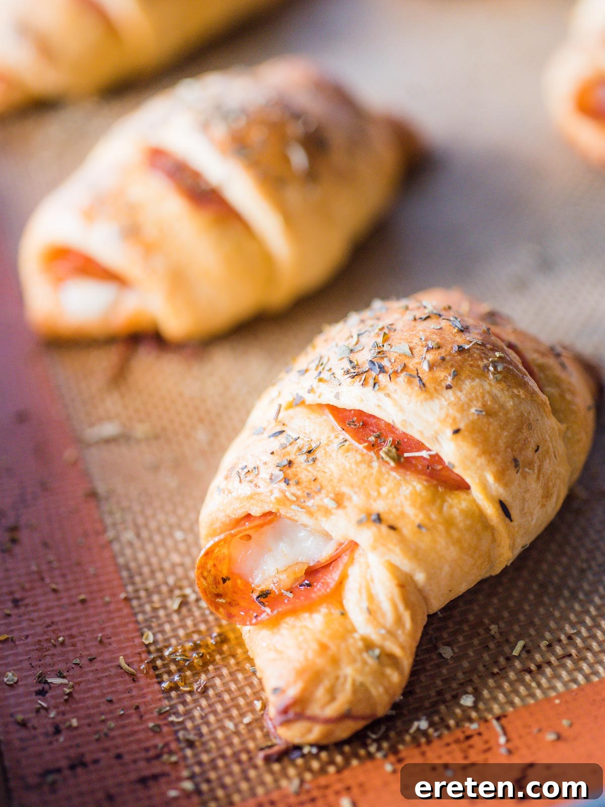 Up close shot of golden brown pepperoni rolls on a silicone-lined baking sheet, showing melted cheese and visible pepperoni slices