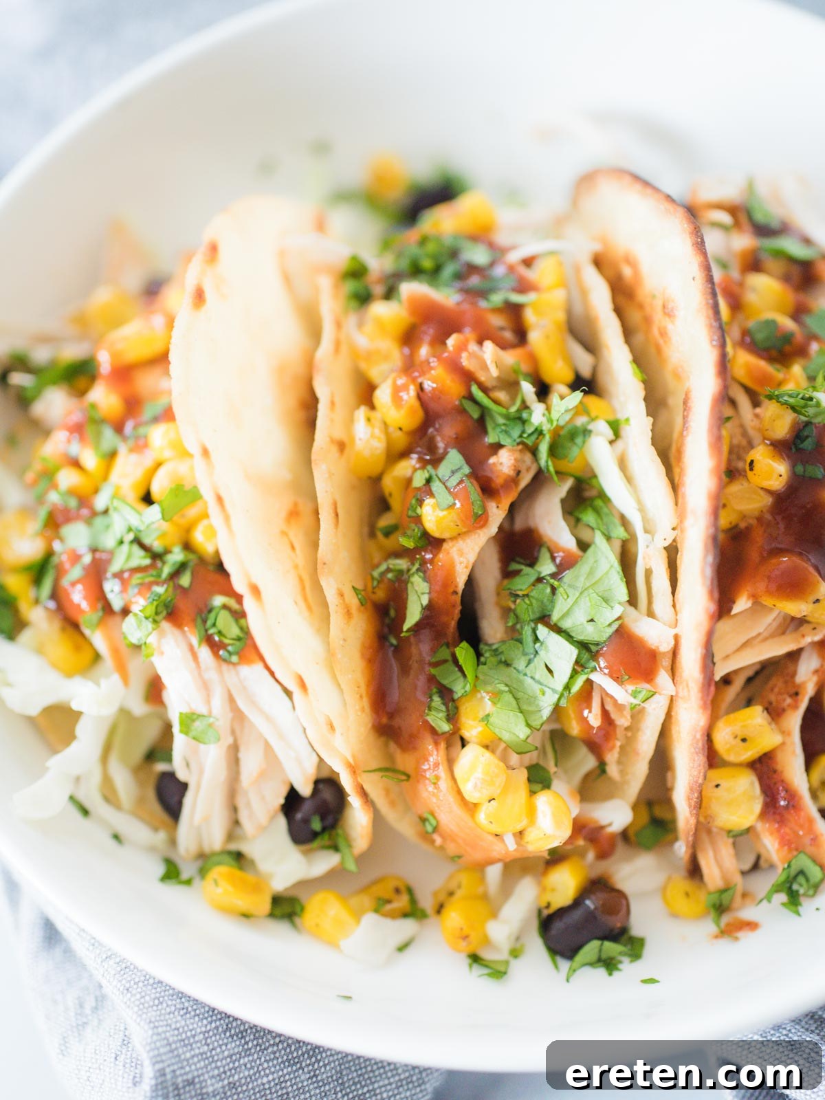 A white plate with two BBQ chicken tacos standing upright, filled with shredded chicken, corn, and other toppings.