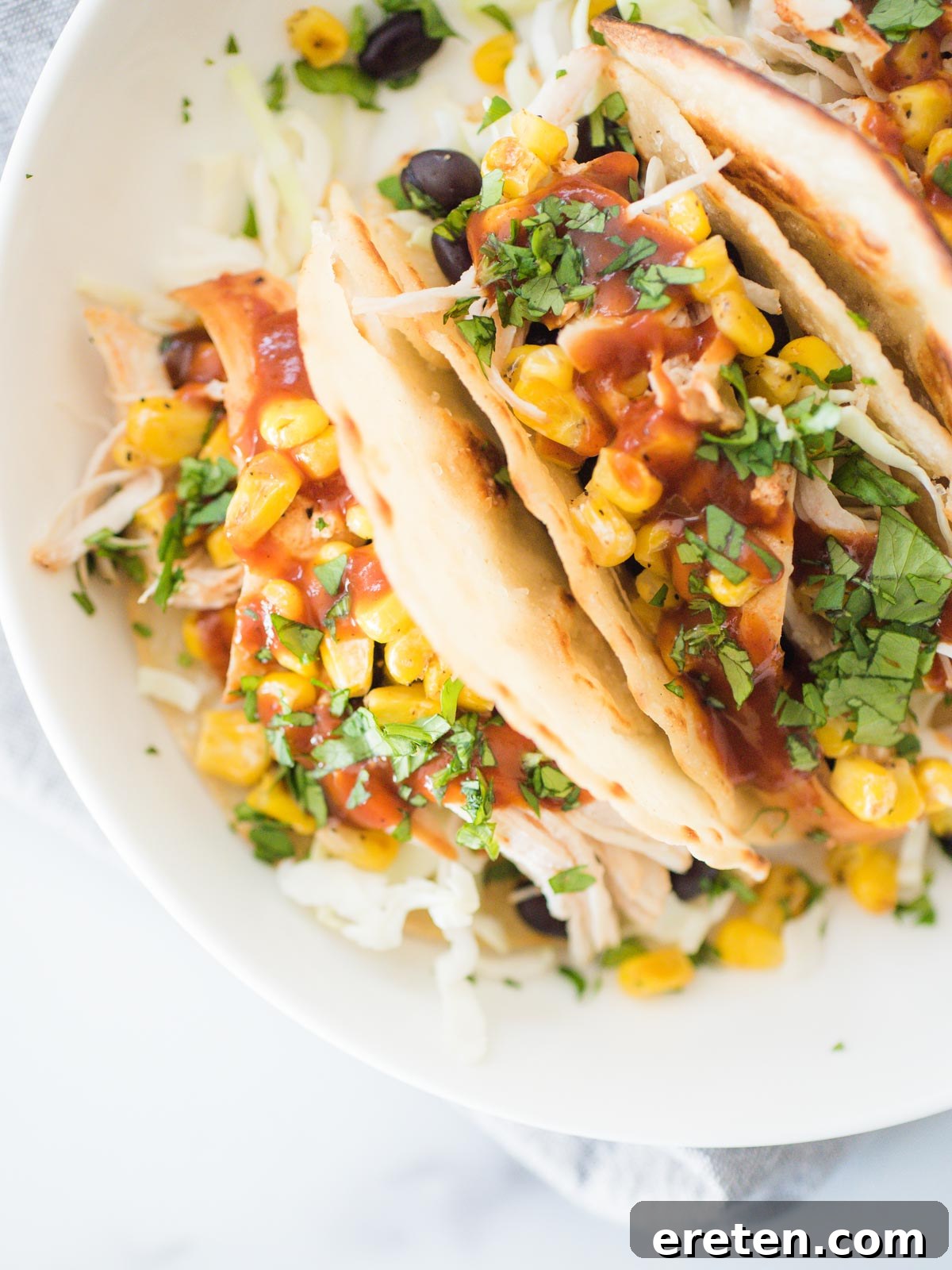 A shallow white bowl filled with three BBQ chicken tacos, garnished with cilantro and served with a side of extra BBQ sauce.