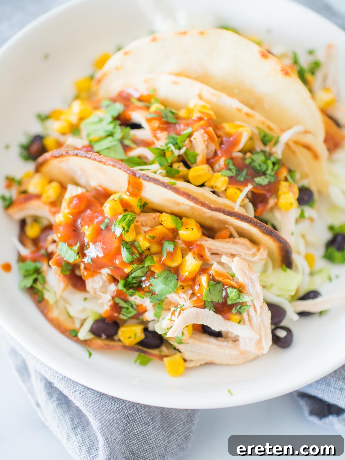 A shallow white bowl presenting two BBQ chicken tacos, generously stuffed with BBQ chicken, black beans, corn, and cilantro, ready to be enjoyed.