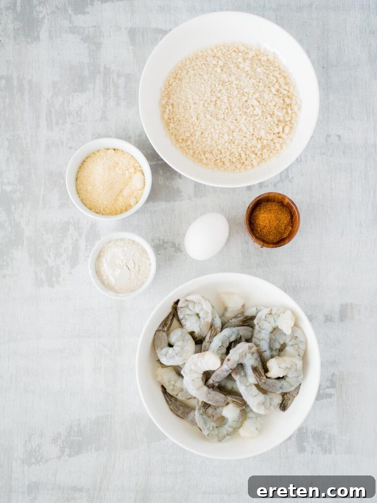 A collection of ingredients laid out, including panko breadcrumbs, Parmesan, Old Bay, flour, eggs, raw shrimp, and avocado oil spray, for making air fryer shrimp.