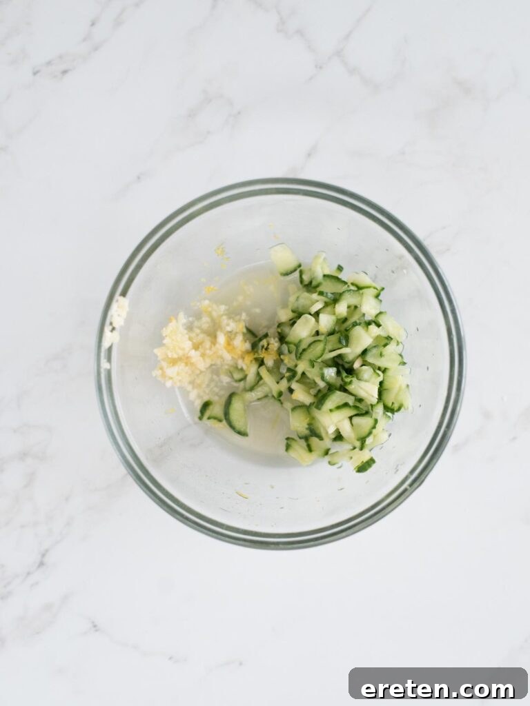 A bowl showcasing the fresh components of tzatziki sauce: English cucumber, fragrant lemon zest, tart lemon juice, and pungent minced garlic.