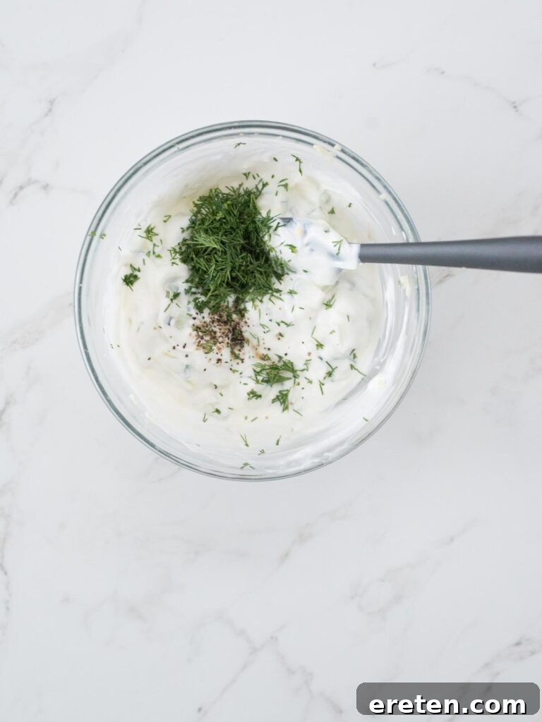 Fresh dill, ground black pepper, and kosher salt are artfully sprinkled over a bowl of creamy Greek yogurt, forming the base of a delicious tzatziki.