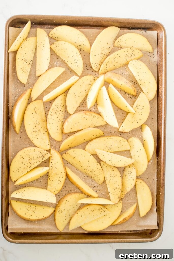 Seasoned potato wedges on a parchment-lined baking sheet, evenly coated with garlic powder, salt, and pepper.