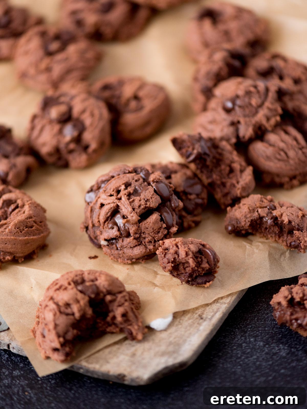 Velvet Triple Chocolate Pudding Cookies 2 A close-up of several chocolate cookies, some whole and some broken, resting on brown parchment paper atop a wooden board. The cookies are studded with chocolate chips.