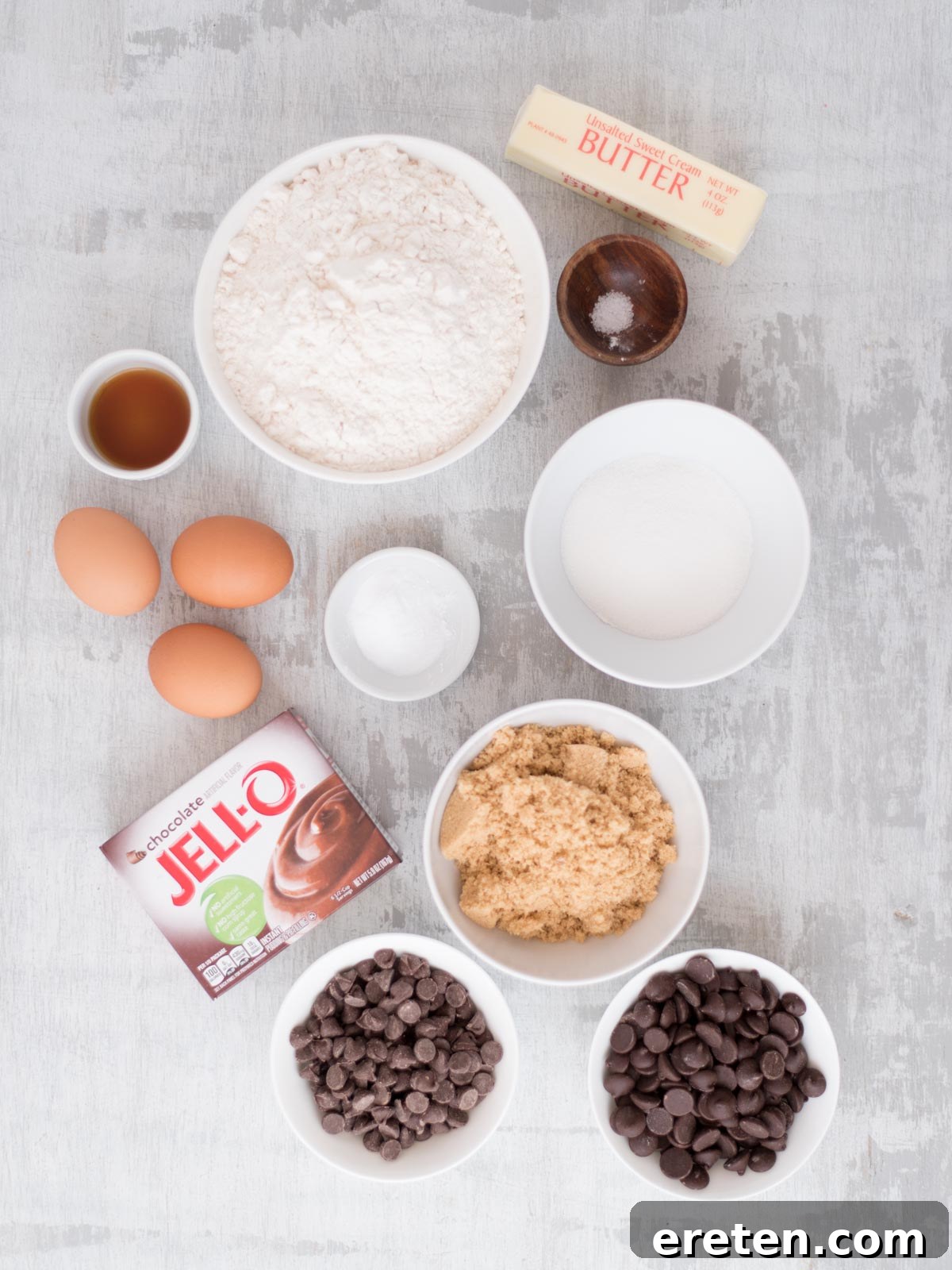 Velvet Triple Chocolate Pudding Cookies 3 A flat lay of baking ingredients includes flour, eggs, butter, vanilla extract, salt, granulated sugar, brown sugar, baking soda, chocolate pudding mix, and two bowls of chocolate chips on a light surface.