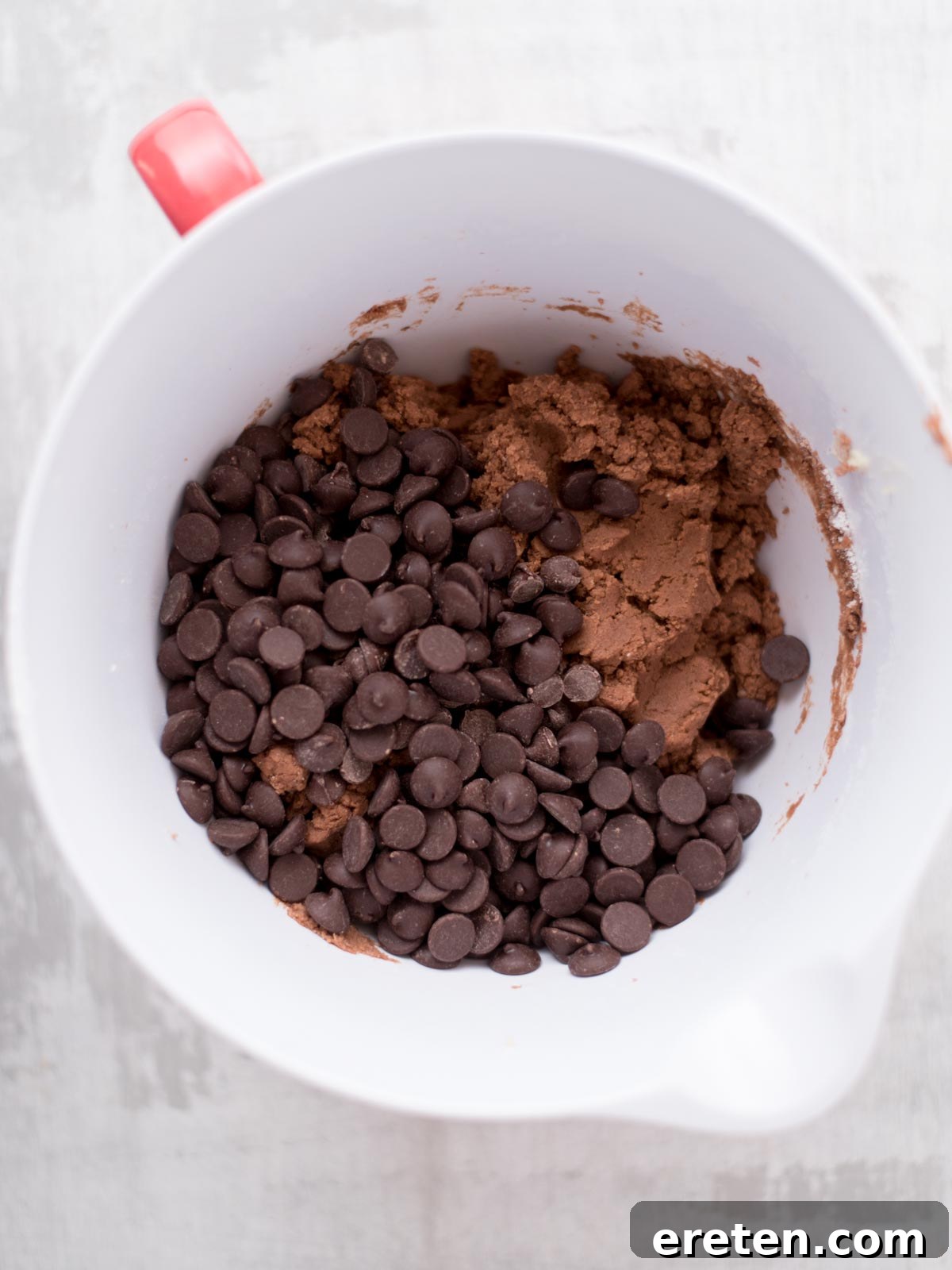 Velvet Triple Chocolate Pudding Cookies 4 A white mixing bowl with a red handle contains chocolate cookie dough and a generous amount of chocolate chips on a light-colored surface.