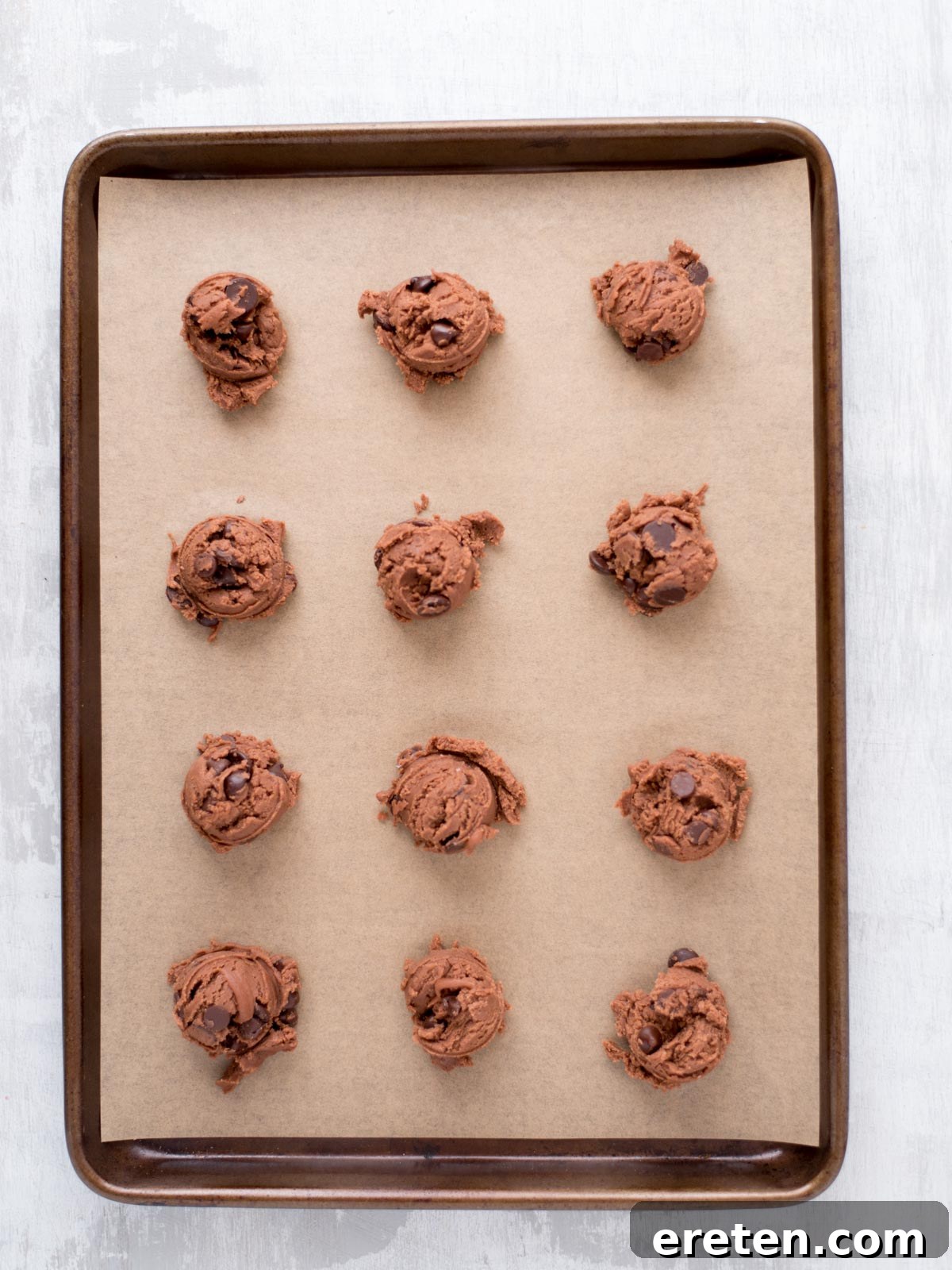Velvet Triple Chocolate Pudding Cookies 5 A baking tray lined with parchment paper holds twelve evenly spaced scoops of chocolate cookie dough, ready to be baked.