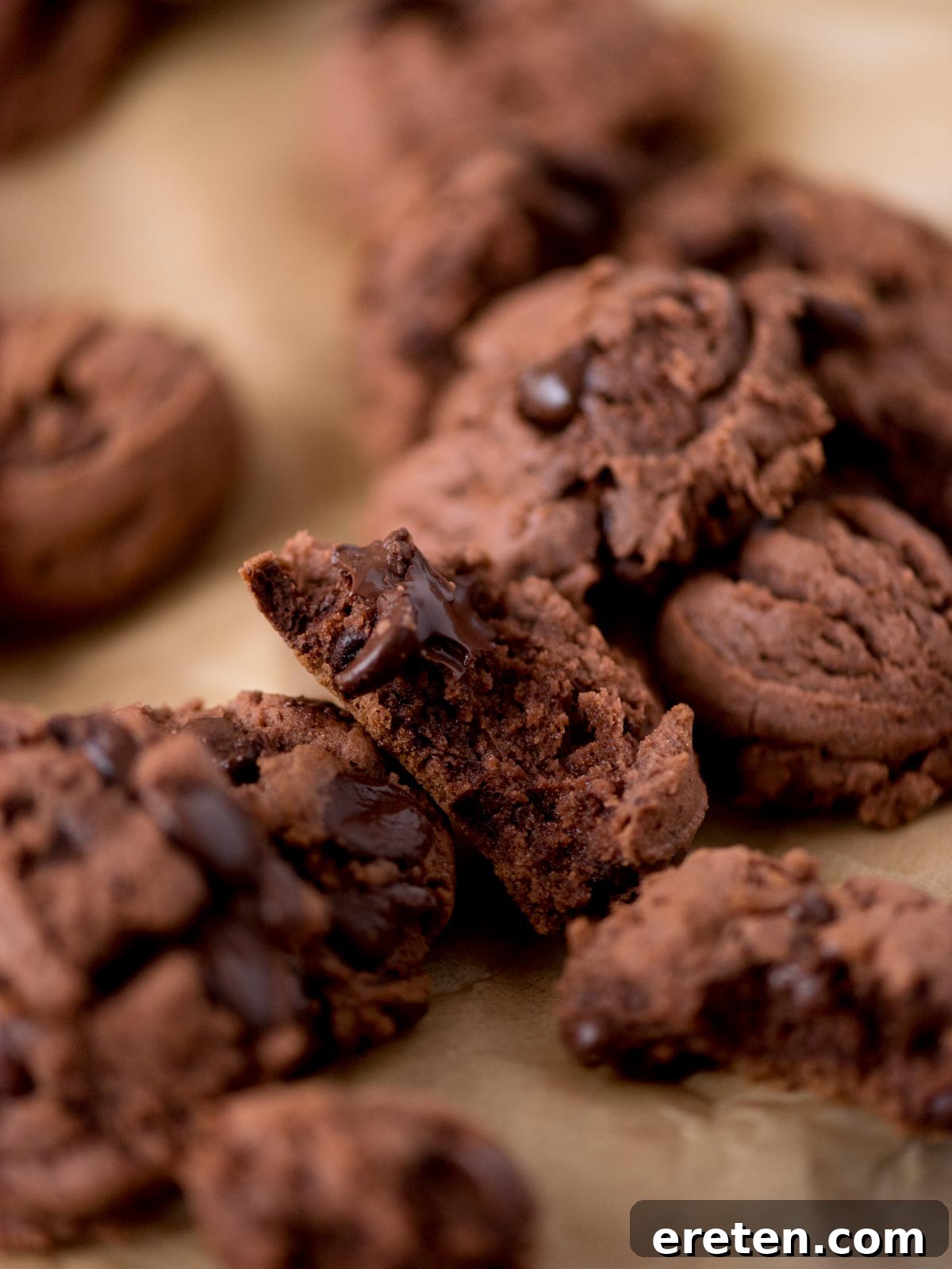 Velvet Triple Chocolate Pudding Cookies 6 A close-up of several chocolate cookies with chocolate chips, some broken in half, resting on a sheet of brown parchment paper.