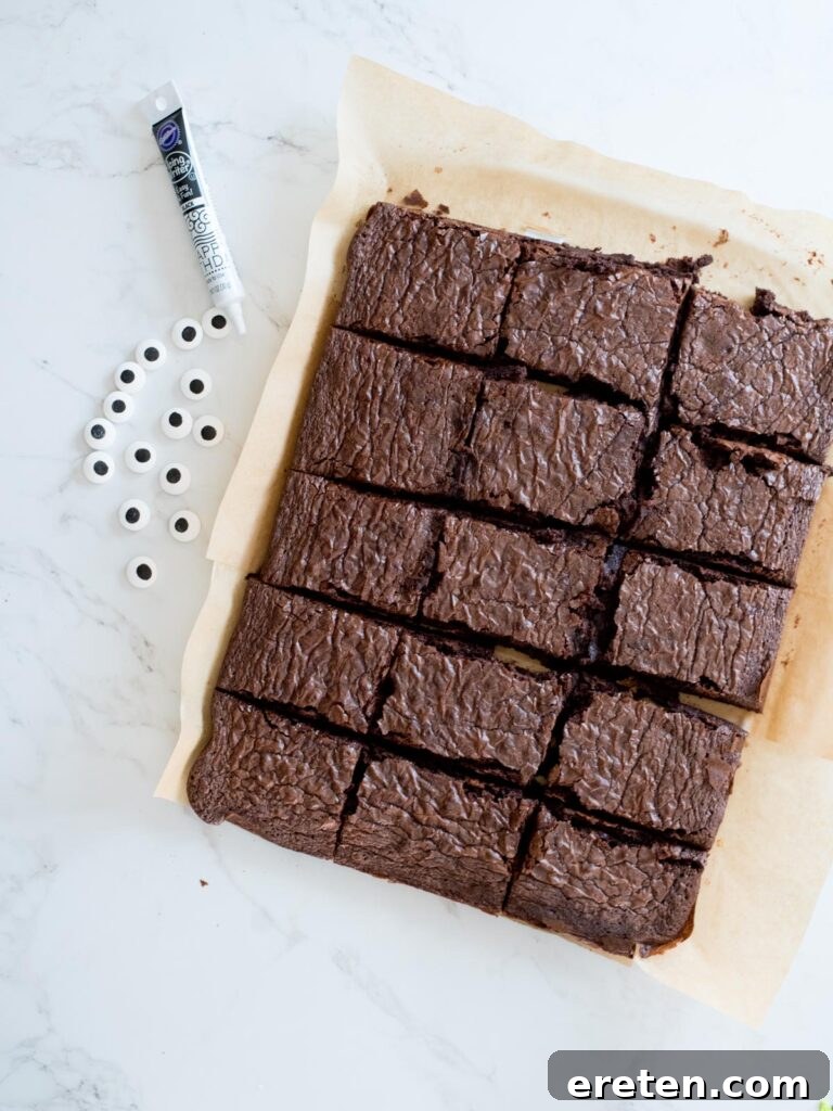 A simple array of Halloween brownie ingredients, including baked brownies, large candy eyes, and a tube of cookie icing, laid out on a clean surface ready for decorating.