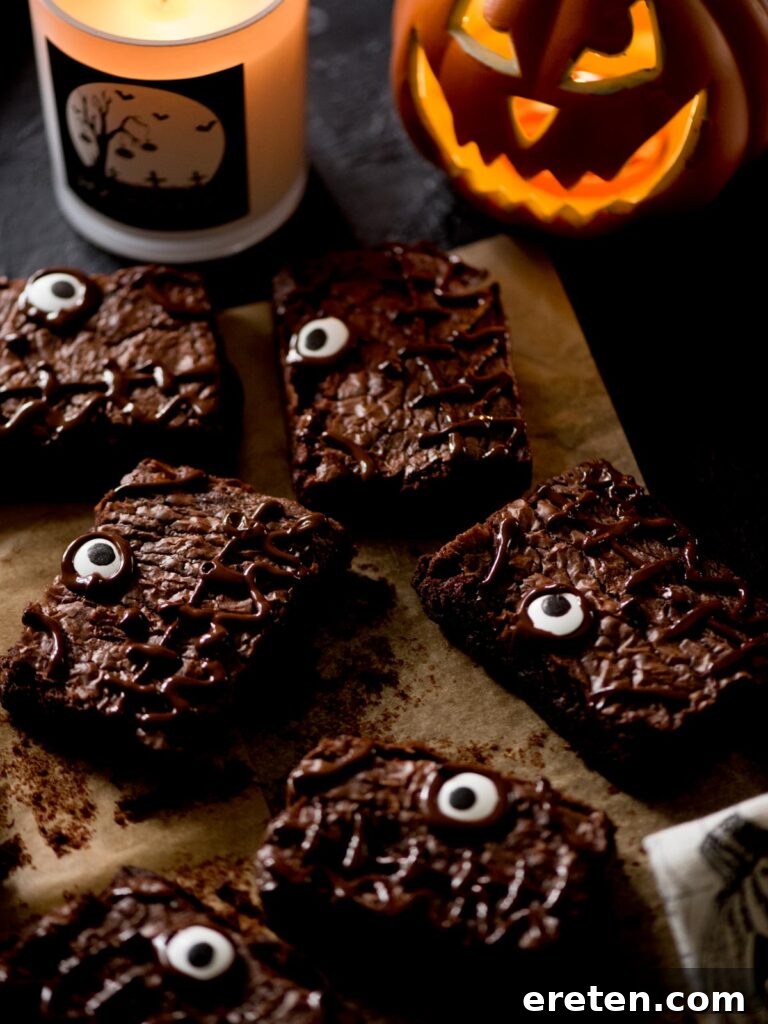 Decorated Halloween brownies with candy eyes, arranged on a rustic wooden board, accompanied by a striped towel, a glowing candle, and a small decorative pumpkin for a festive ambiance.