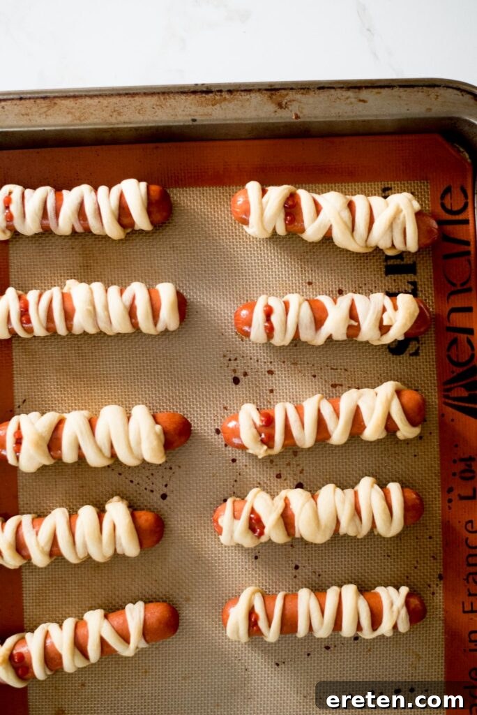 Halloweenie Mummies 5 Mummy pigs in a blanket arranged on a baking sheet before being placed in the oven