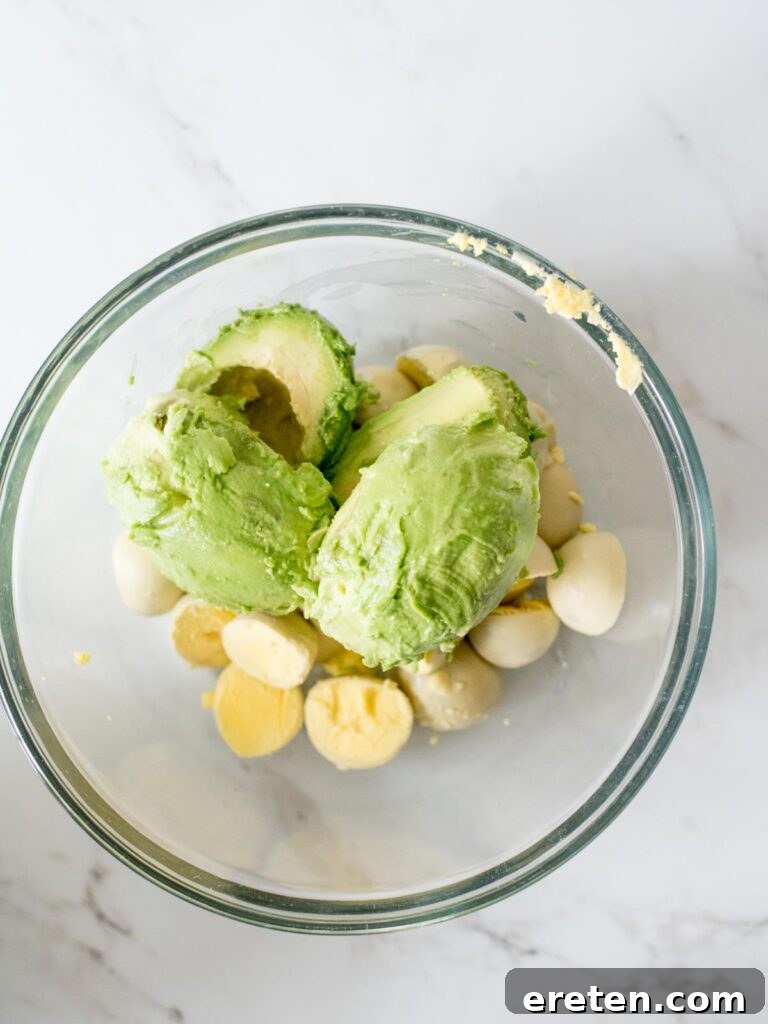 Sliced avocado being added to the mixing bowl containing the mashed hard-boiled egg yolks.