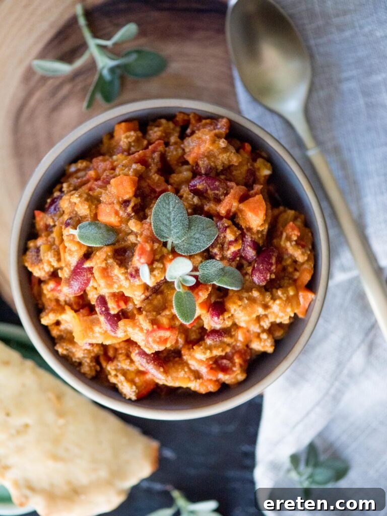 Hearty pumpkin chili served in a white bowl, adorned with a sprig of fresh sage, inviting a warm, comforting meal.
