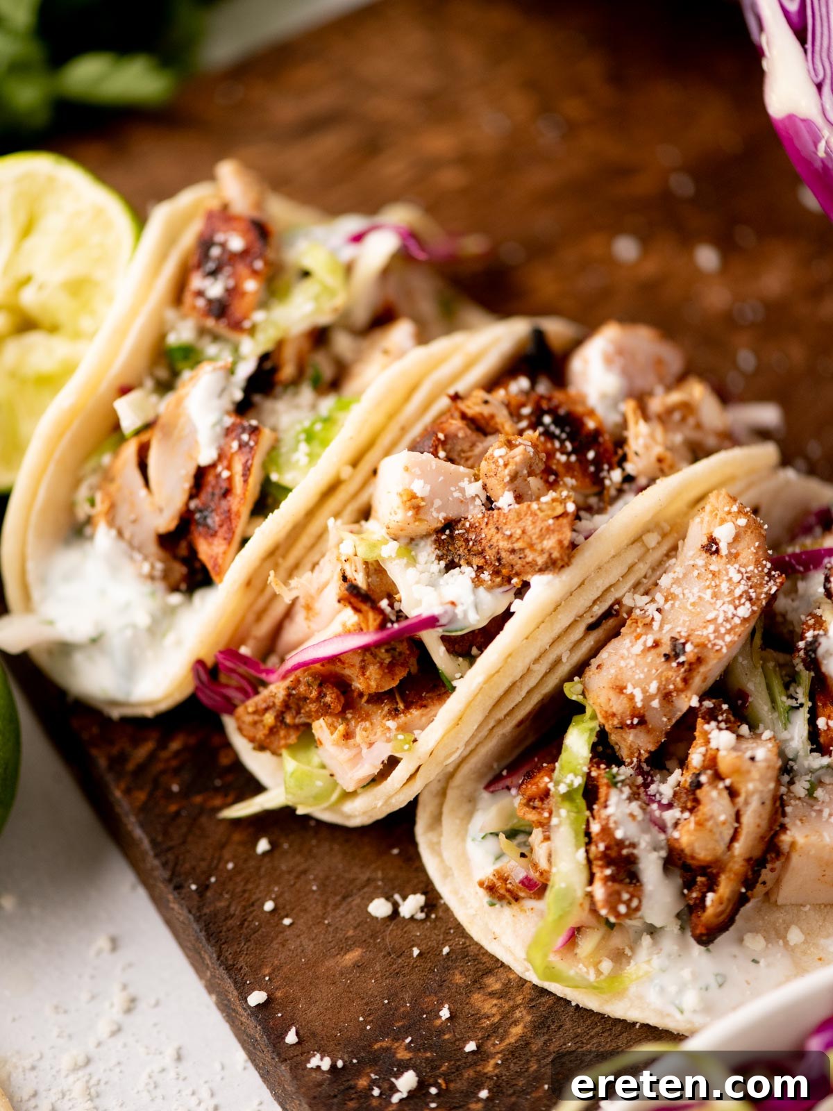 Zesty Street Chicken Tacos 2 Three soft tacos filled with grilled chicken, shredded lettuce, red cabbage, creamy sauce, and crumbled cheese are arranged on a wooden board. A halved lime sits nearby.