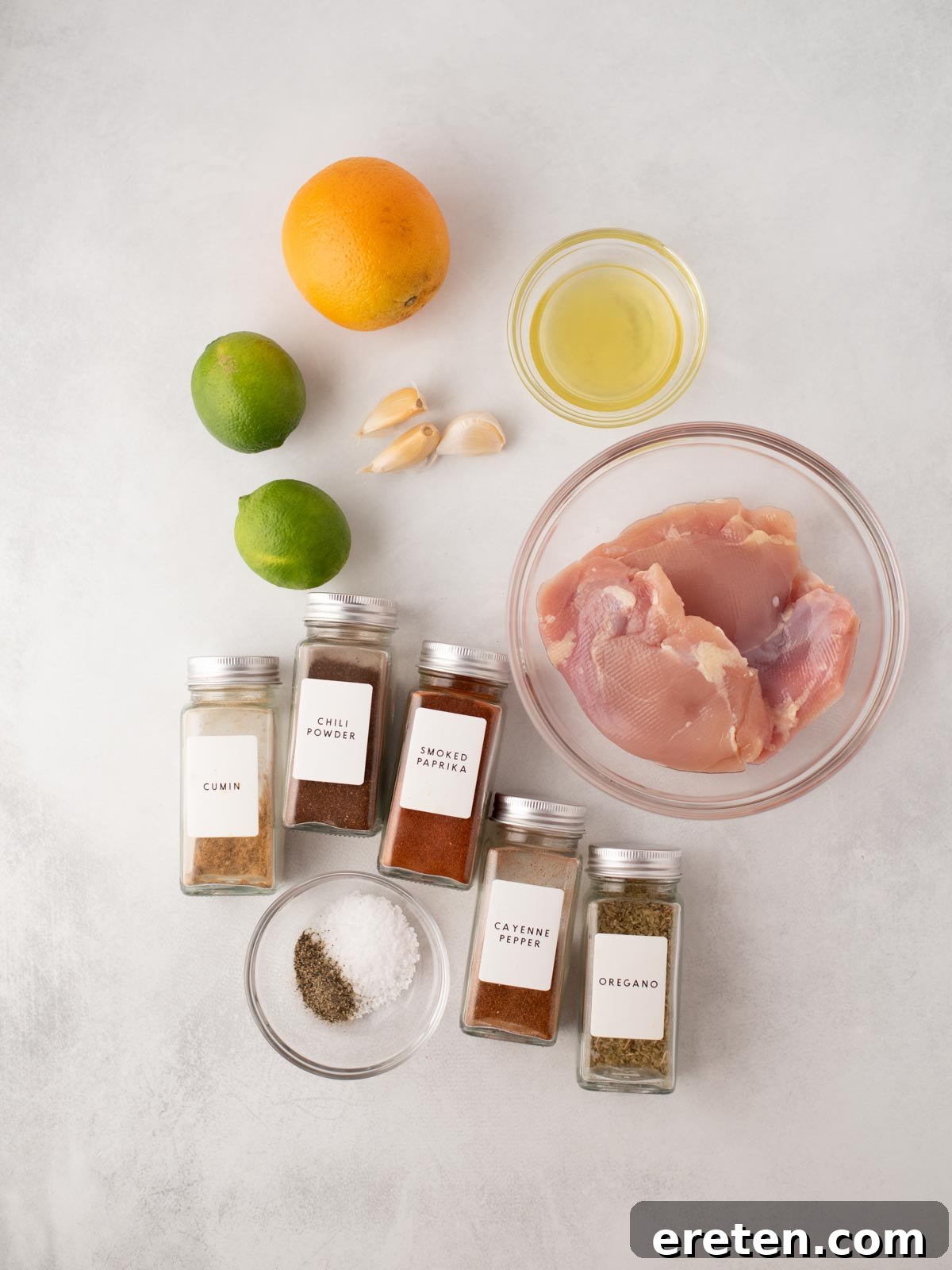 Zesty Street Chicken Tacos 3 Raw chicken breast in a glass bowl, surrounded by spices in jars (cumin, chili powder, smoked paprika, cayenne pepper, oregano), black pepper, salt, garlic, lime, orange, and a small bowl of oil on a white surface.