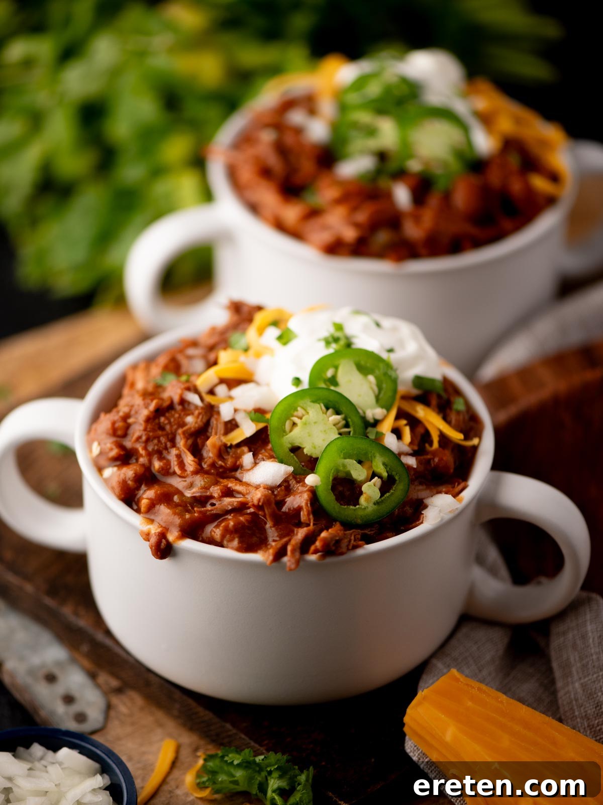 Tender Chuck Roast Chili 2 Two white bowls filled with chili, topped with sliced jalapeños, shredded cheese, and sour cream, sit on a wooden board. Fresh herbs and cheese are visible in the background, hinting at fresh ingredients.