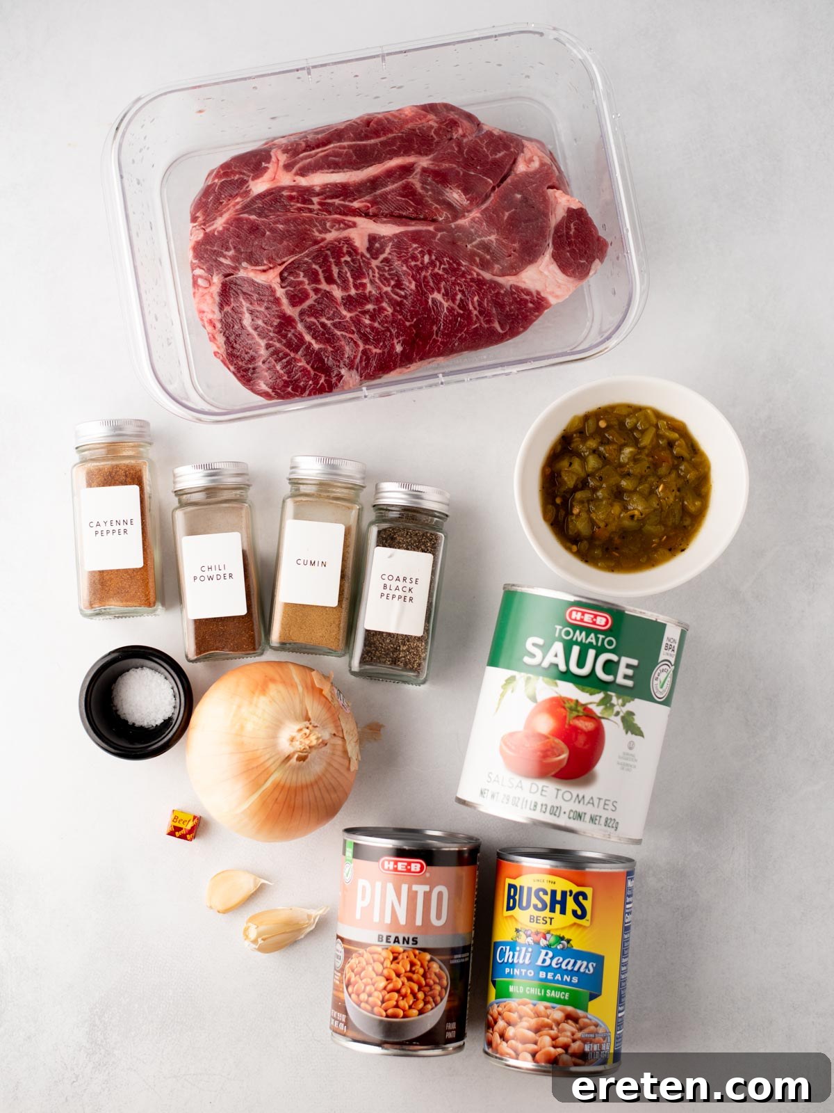 Tender Chuck Roast Chili 3 Ingredients for chili are arranged on a light surface, including a large cut of raw beef, cans of tomato sauce, pinto beans, and chili beans, spices in jars, an onion, garlic cloves, a bouillon cube, and a bowl of green chilies, all ready for cooking.