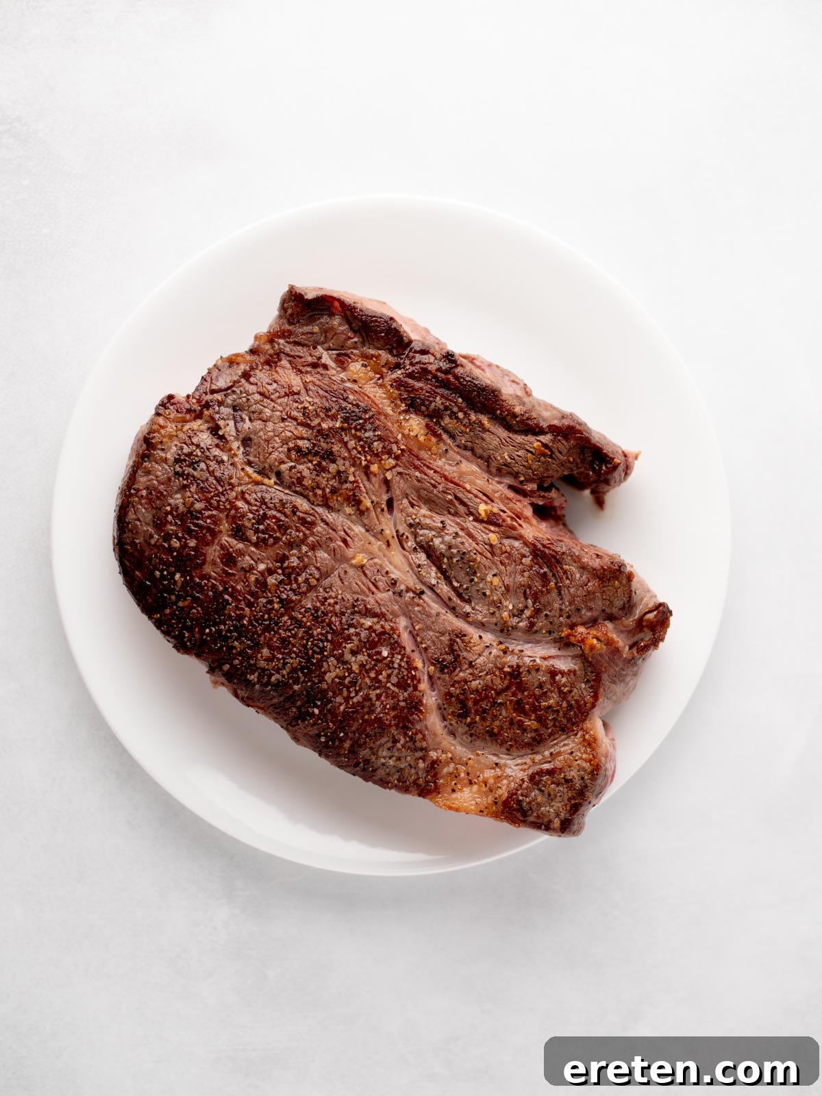Tender Chuck Roast Chili 4 A perfectly seared, seasoned chuck roast sits on a white plate against a light background. The beef has a rich, browned exterior with visible spices, indicating a well-developed crust.