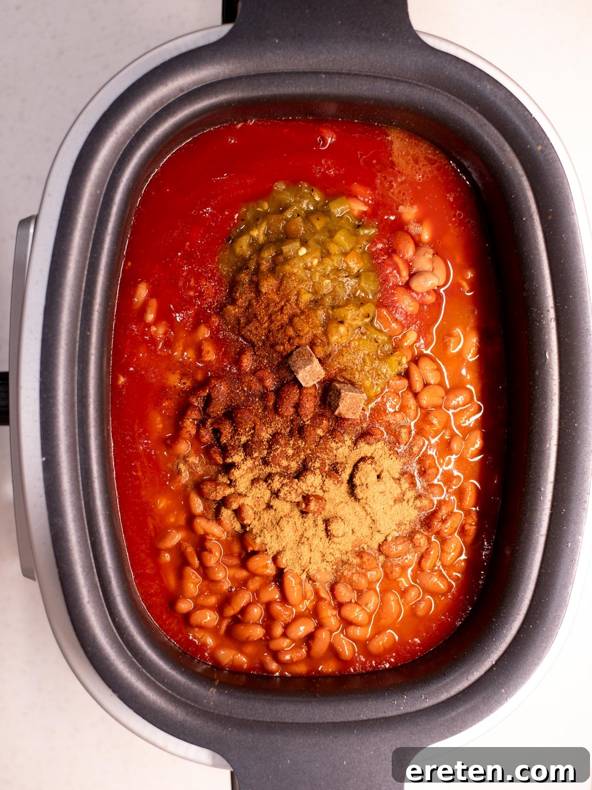 Tender Chuck Roast Chili 5 Overhead view of a slow cooker filled with a rich base of tomato sauce, various spices, chopped green chilies, and both pinto and chili beans, layered over the seared beef roast, ready for mixing and slow cooking.