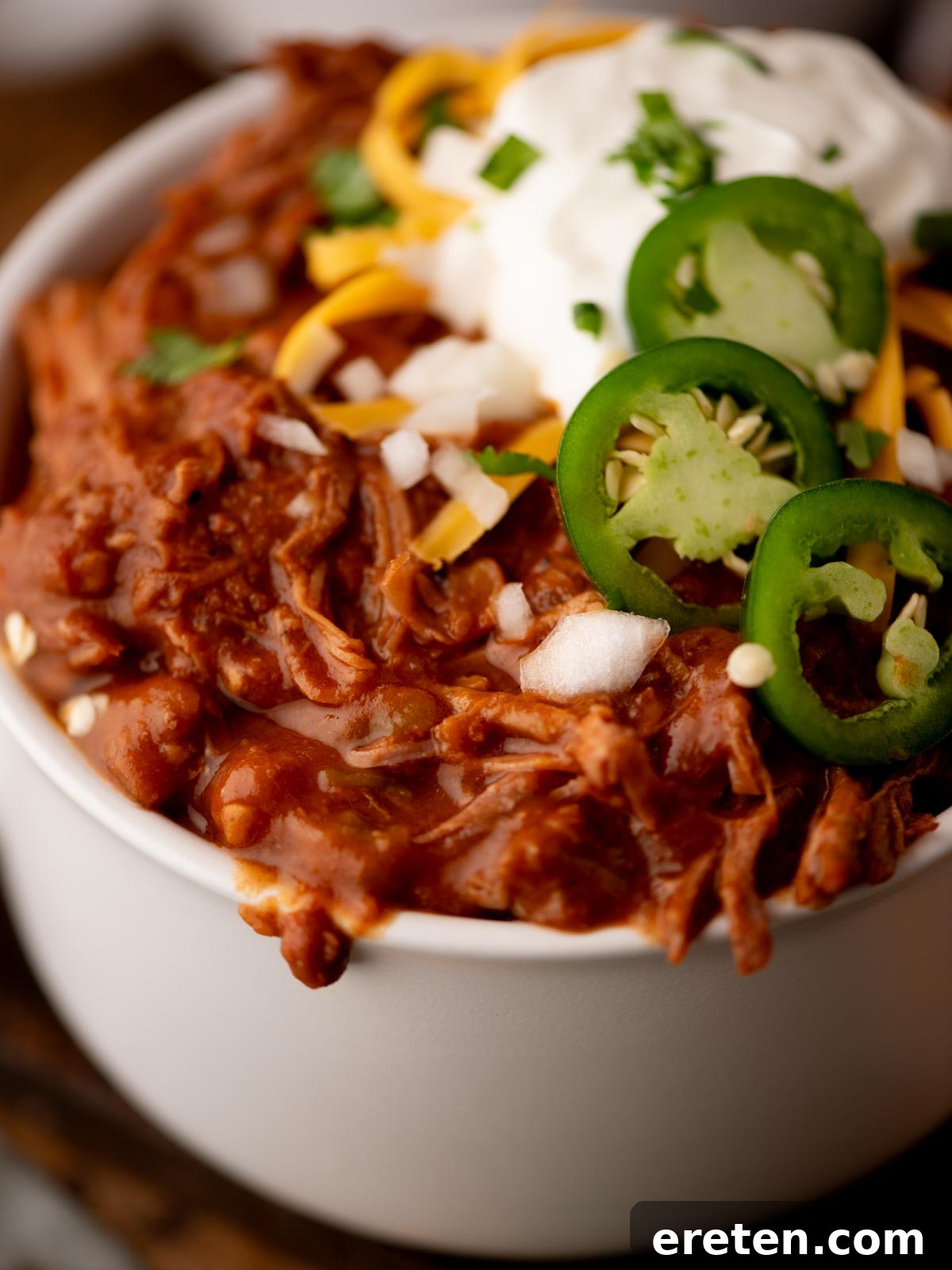 Tender Chuck Roast Chili 6 A vibrant bowl of chili generously topped with thinly sliced jalapeños, a heap of shredded cheddar cheese, a dollop of cool sour cream, finely chopped red onions, and fresh cilantro sprigs. The chili itself appears thick, rich, and hearty, showcasing tender shredded meat within its robust sauce.