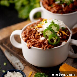 Tender Chuck Roast Chili 7 A white bowl of chili topped with sliced jalapenos, shredded cheese, chopped onions, and a dollop of sour cream, sits on a wooden board with fresh herbs and a block of cheddar cheese nearby.
