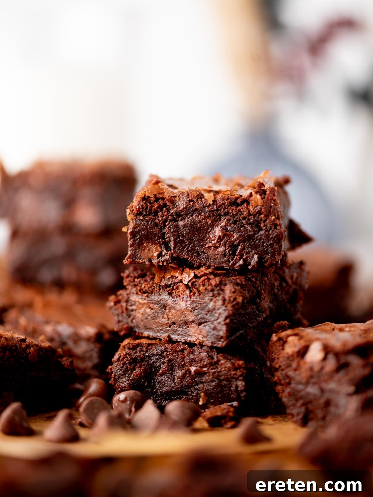 A close-up of a stack of rich, fudgy chocolate brownies with a bite taken out of the top brownie, surrounded by chocolate chips, with more brownies blurred in the background, highlighting the desirable crackly top and moist interior.