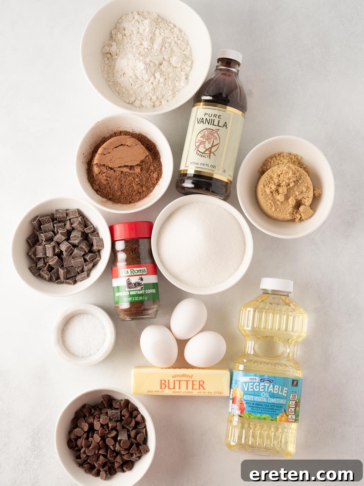 Assorted baking ingredients on a white surface: flour, vanilla extract, brown and white sugar, cocoa powder, instant coffee, chocolate chunks, chocolate chips, eggs, butter, vegetable oil, and salt, ready for brownie preparation.