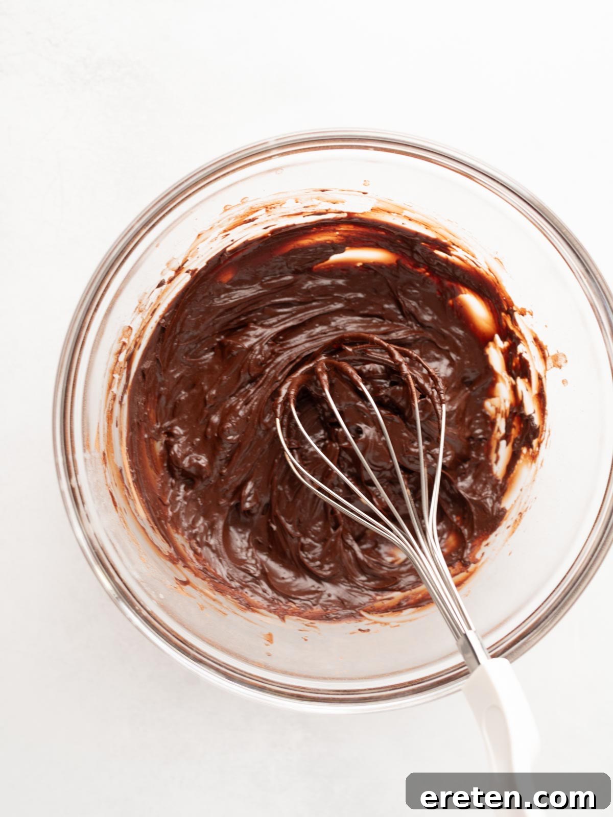 A glass bowl filled with thick chocolate batter being mixed with a metal whisk on a white surface, demonstrating the initial cocoa blooming step.
