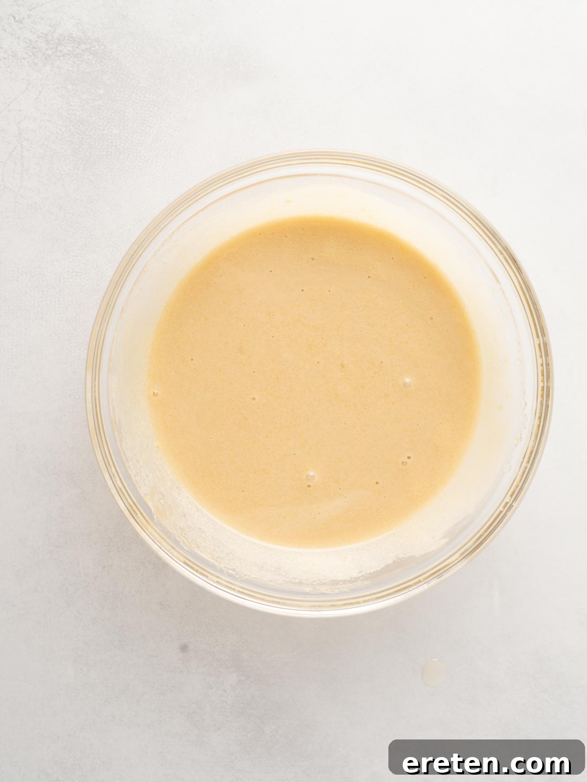 A clear glass bowl filled with a smooth, light tan batter, the result of whisking eggs and sugars until airy and glossy, sits on a white surface.