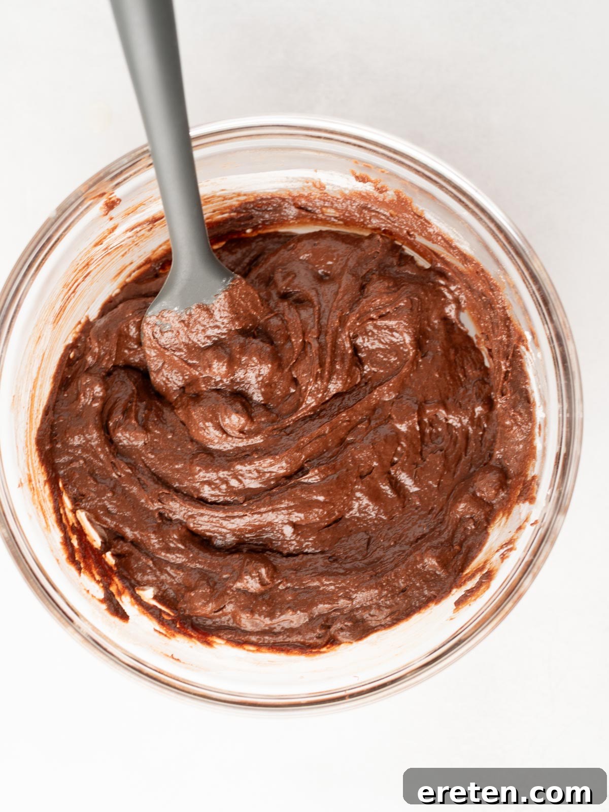 A glass bowl filled with thick, swirled chocolate brownie batter, with a spoon resting inside. Chocolate chunks are visible throughout the rich, dark batter, ready for baking.