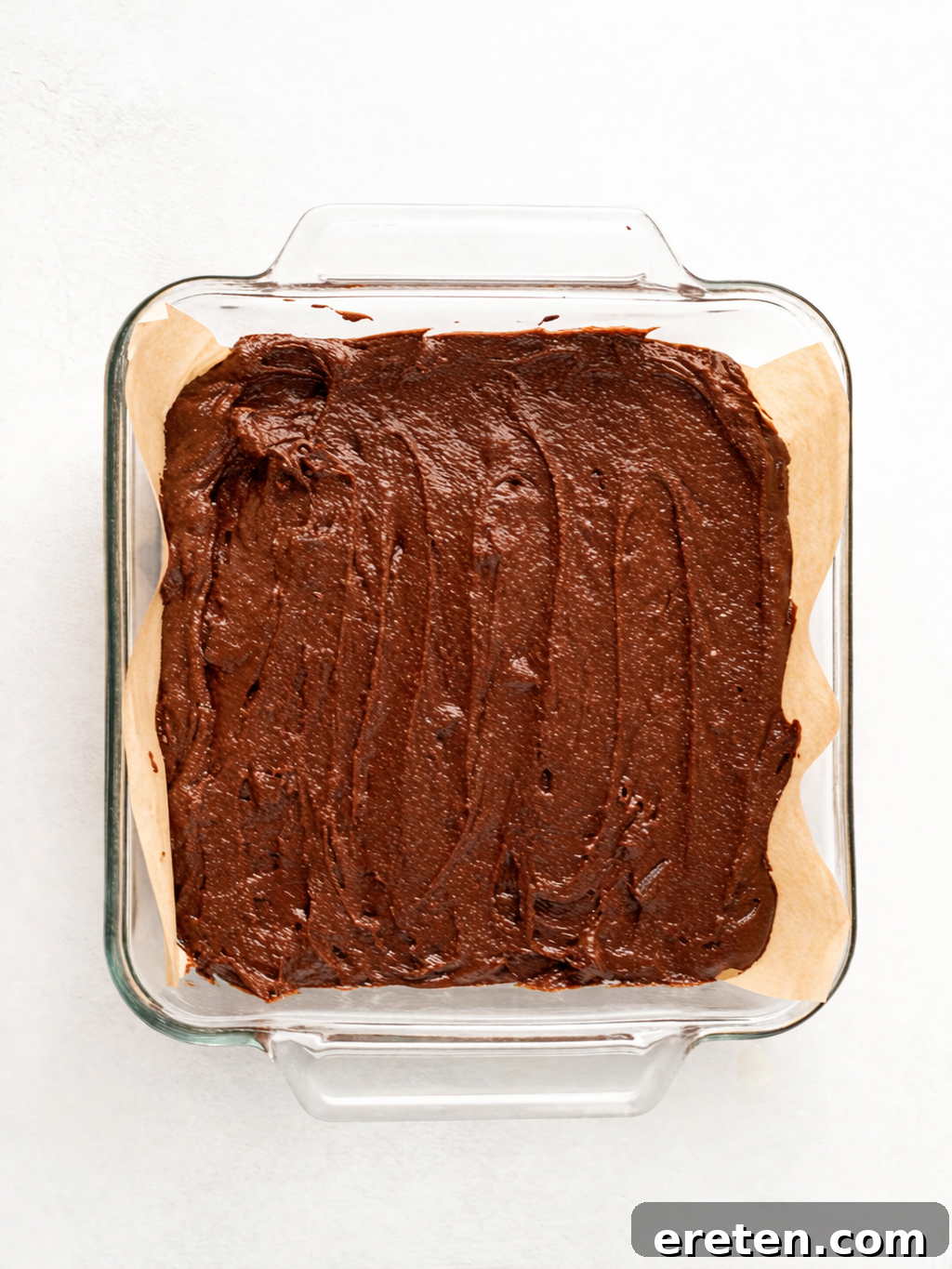 A glass baking dish lined with parchment paper is filled with unbaked chocolate brownie batter, spread evenly across the surface, ready to go into the oven.