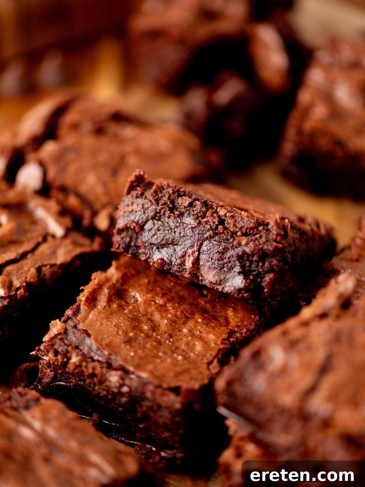 Close-up of rich, fudgy chocolate brownies with a crackly top, stacked and surrounded by other brownie pieces, showcasing their moist, dense texture and appealing finish.