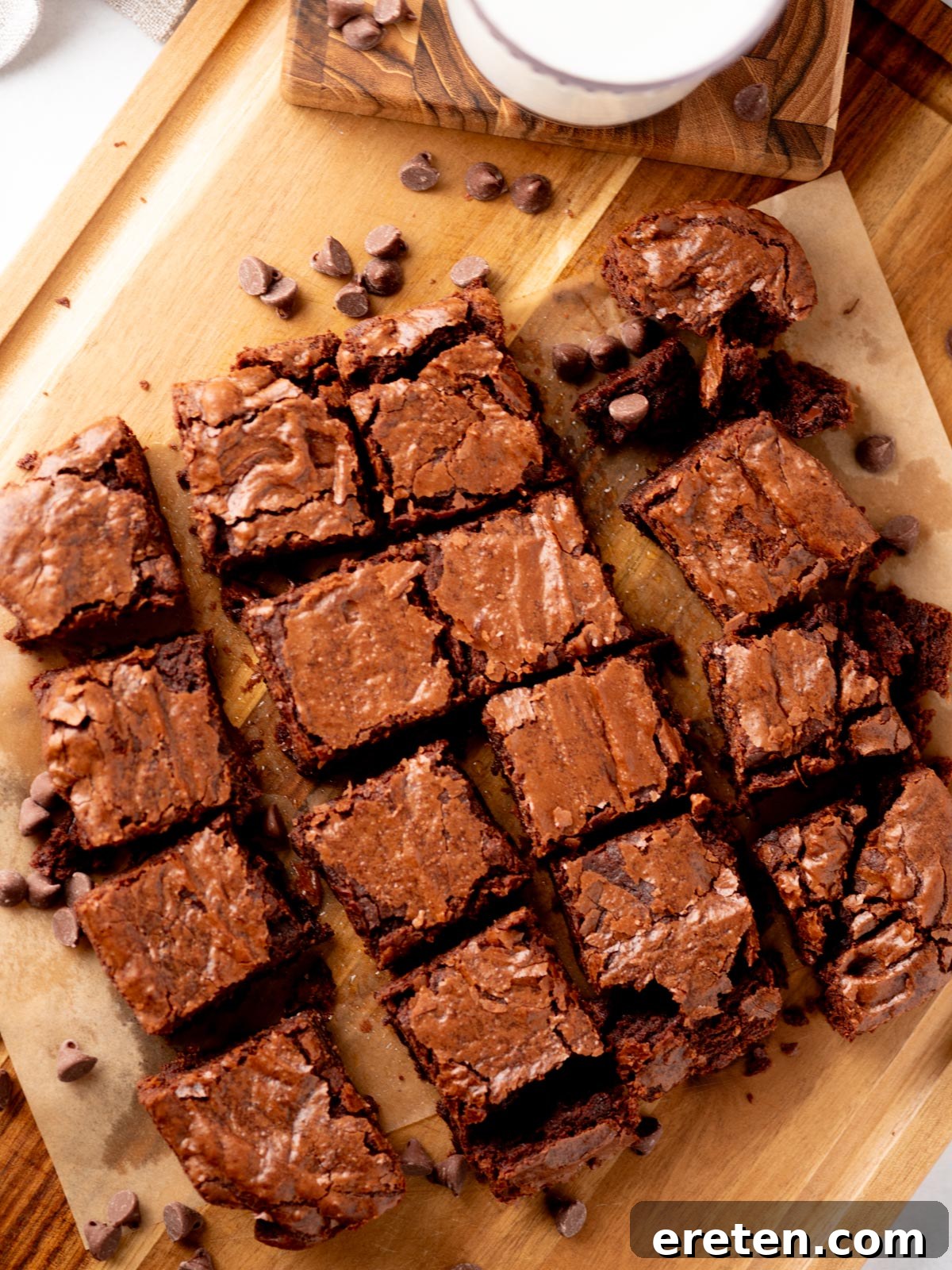 Freshly baked chocolate brownies cut into squares are arranged on parchment paper atop a wooden board, surrounded by scattered chocolate chips. A tall glass of milk sits nearby, inviting a delightful pairing.