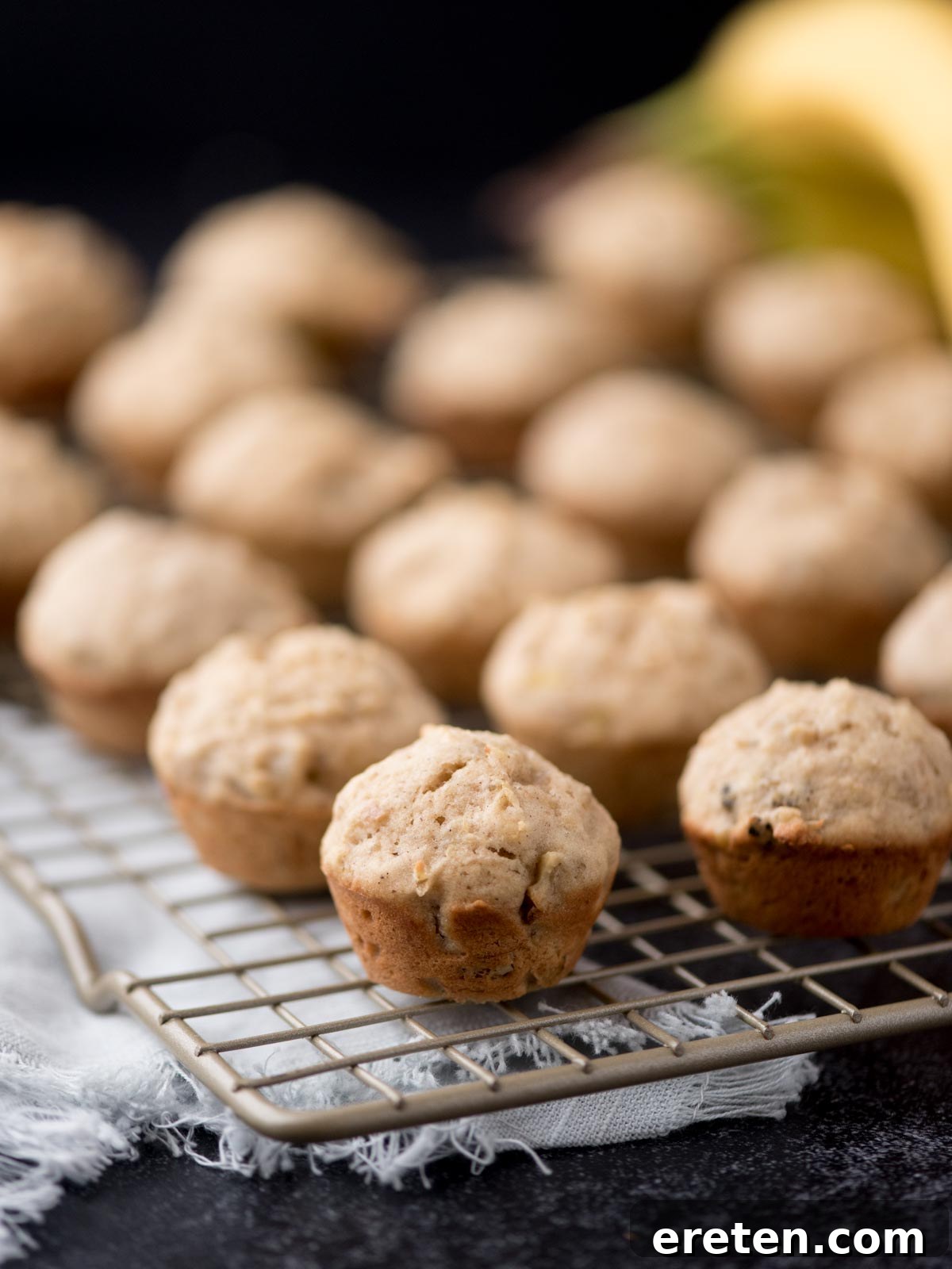 A wire cooling rack filled with freshly baked mini banana nut muffins, showcasing their golden-brown tops and fluffy texture. Several muffins in the foreground are in sharp focus.