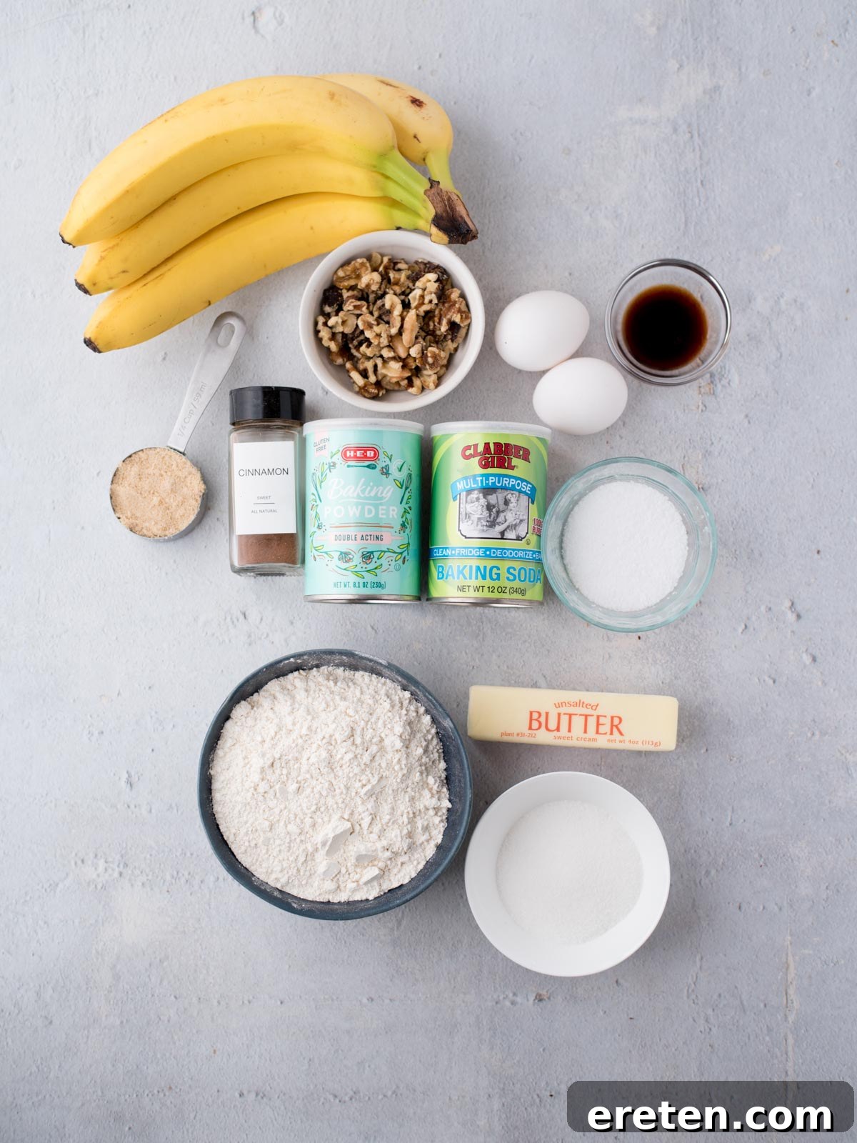 An overhead view of all the ingredients for mini banana nut muffins arranged on a countertop, including flour, sugars, butter, eggs, overripe bananas, and walnuts.
