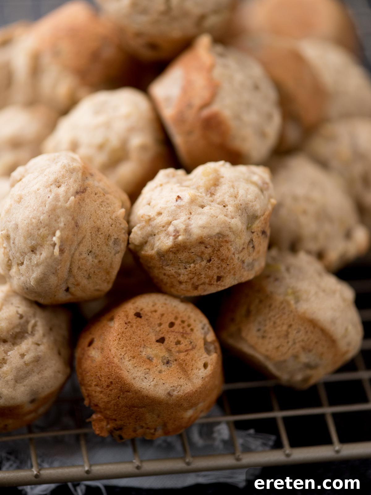 A close-up photograph of several mini banana nut muffins stacked on a wire cooling rack, highlighting their perfectly golden-brown color and soft texture.