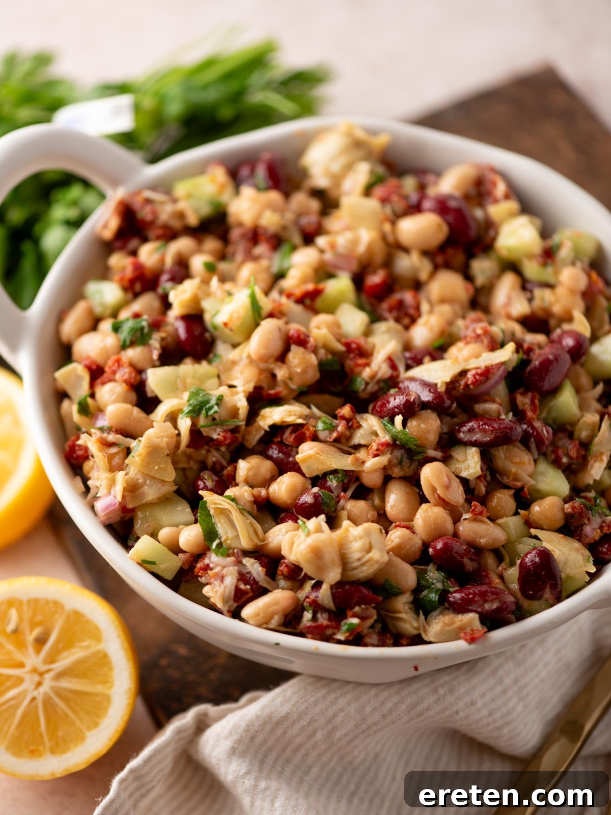 A white bowl filled with a colorful bean salad containing kidney beans, chickpeas, artichoke hearts, chopped cucumbers, herbs, and sun-dried tomatoes, with lemon halves and fresh parsley nearby on a wooden surface.