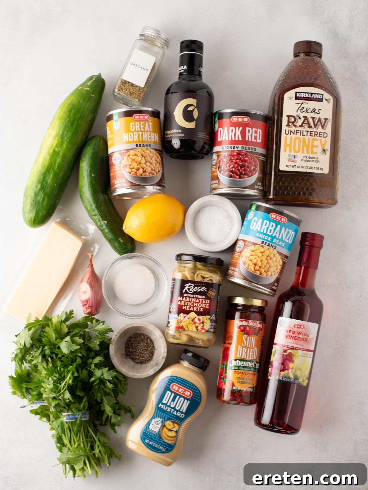 A flat lay of various ingredients, including canned beans, cucumber, lemon, honey, red wine vinegar, Dijon mustard, herbs, sun-dried tomatoes, cheese, black pepper, salt, olives, and a small bottle of balsamic vinegar.