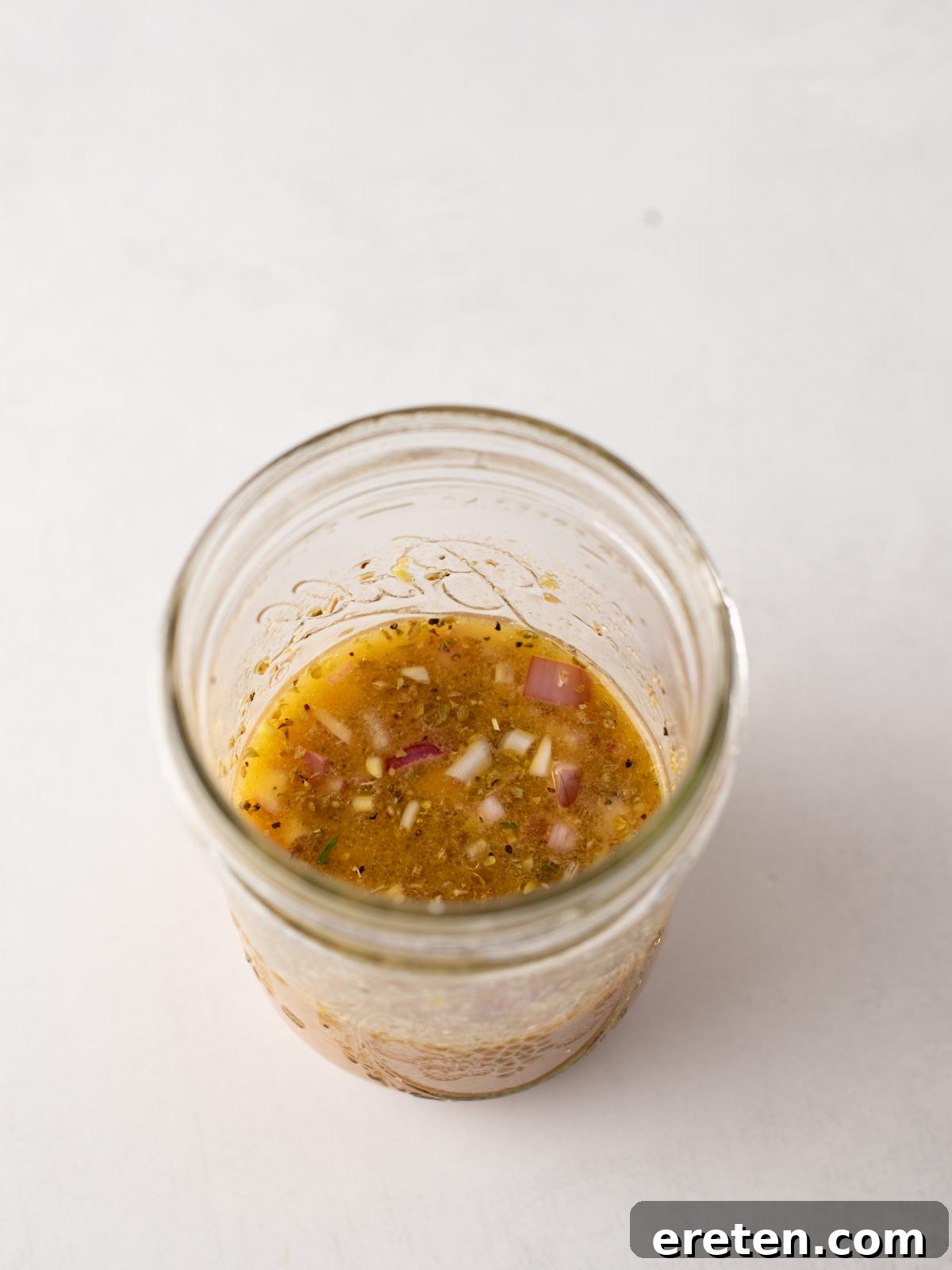 A glass jar filled with a chunky vinaigrette-style dressing containing visible bits of red onion and herbs, set against a plain white background.