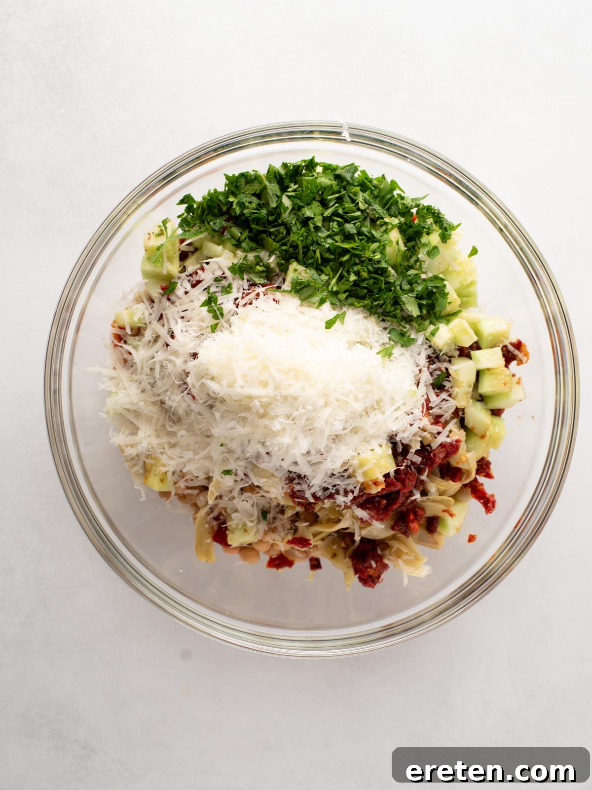 A glass bowl filled with chopped parsley, grated cheese, diced celery, sun-dried tomatoes, and artichoke hearts on a white background.